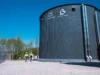 Finlandia inaugura la batería de arena más grande del mundo en Pornainen Dos personas con chalecos reflectantes y cascos caminan junto a un gran silo cilíndrico gris oscuro de la empresa Polar Night Energy y Lovisan Lämpö, bajo un cielo despejado, rodeado de árboles y estructuras industriales.