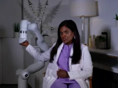 Tampa estrena el primer masaje robótico con inteligencia artificial en la región Una mujer con bata blanca y blusa morada está sentada mientras sostiene el extremo de un brazo robótico blanco. El entorno es moderno y elegante, con plantas, lámparas y decoración de oficina al fondo.