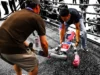 China celebra los primeros Juegos Olímpicos de Robots con resultados hilarantemente caóticos Un pequeño robot humanoide con guantes de boxeo siendo asistido tras caer en lo que parece un ring de combate, mientras varias personas lo observan y fotografían desde el exterior. La escena, capturada con un efecto visual que resalta en color solo al robot y a sus cuidadores, refleja de forma simbólica los desafíos actuales en el desarrollo de la robótica, donde el espectáculo y la innovación se cruzan con la fragilidad de la tecnología en evolución.