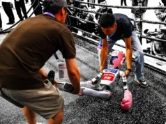 China celebra los primeros Juegos Olímpicos de Robots con resultados hilarantemente caóticos Un pequeño robot humanoide con guantes de boxeo siendo asistido tras caer en lo que parece un ring de combate, mientras varias personas lo observan y fotografían desde el exterior. La escena, capturada con un efecto visual que resalta en color solo al robot y a sus cuidadores, refleja de forma simbólica los desafíos actuales en el desarrollo de la robótica, donde el espectáculo y la innovación se cruzan con la fragilidad de la tecnología en evolución.