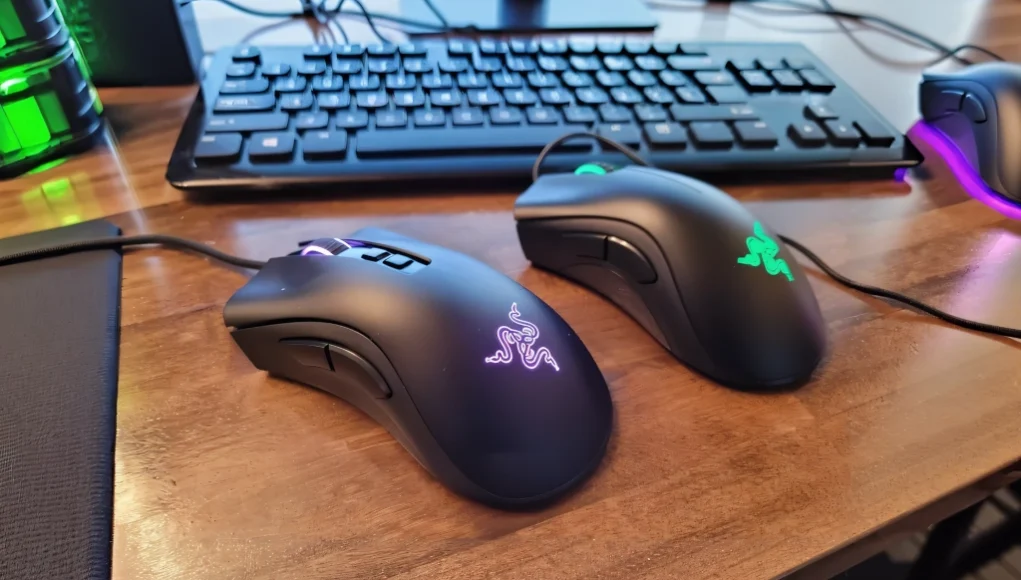 Dos ratones para gaming de la marca Razer sobre un escritorio de madera. Ambos tienen el logotipo iluminado, uno en color púrpura y el otro en verde. Al fondo se ve parcialmente un teclado negro.