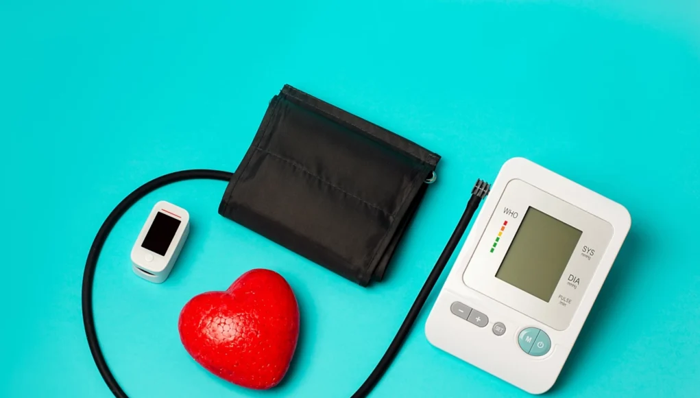 Equipo médico sobre fondo azul: un tensiómetro digital con brazalete negro, un oxímetro de pulso blanco y un corazón rojo decorativo, simbolizando el monitoreo de la salud cardiovascular.