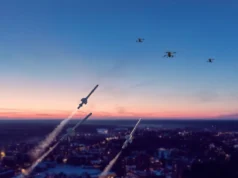 Saab presenta el misil compacto Nimbrix para neutralizar enjambres de drones Ilustración de un cielo al atardecer sobre una ciudad donde varios misiles son lanzados hacia un grupo de drones en vuelo.