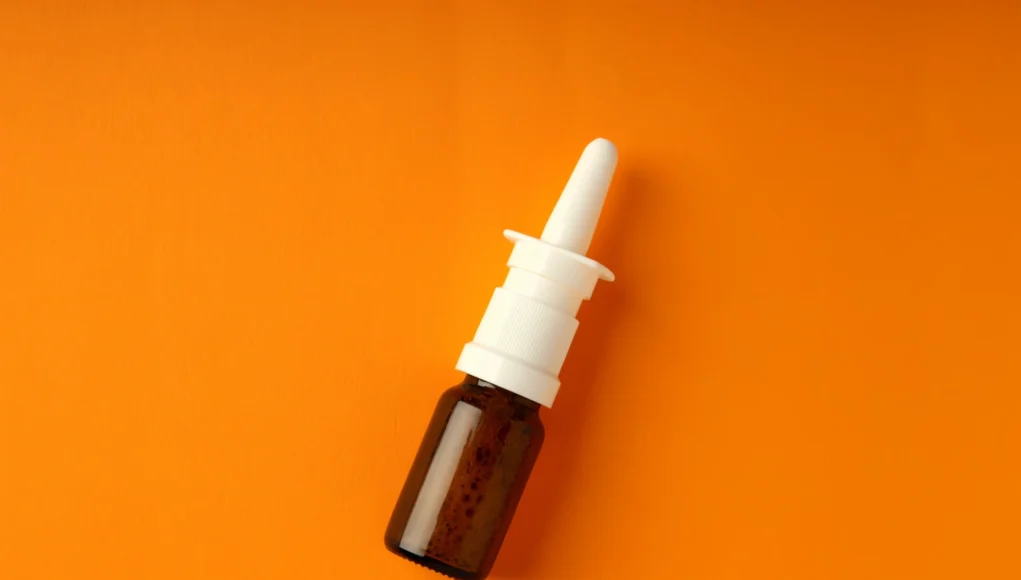 Frasco marrón pequeño con aplicador en spray nasal blanco, colocado sobre un fondo naranja.