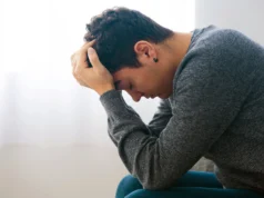 Nueva terapia de estimulación cerebral precisa demuestra eficacia rápida contra la depresión Joven sentado con la cabeza inclinada hacia adelante y las manos sobre el cabello, mostrando una expresión de angustia o preocupación.