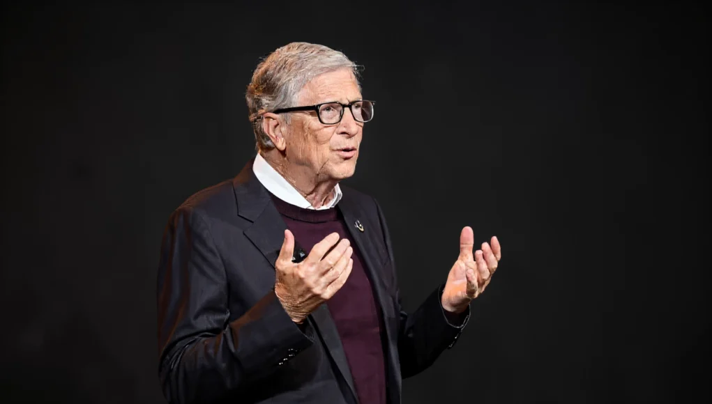 Bill Gates IA Empleo Hombre mayor con gafas y traje oscuro hablando en un escenario, gesticulando con las manos frente a un fondo negro.