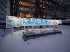 La batería de cemento del MIT acaba de volverse 10 veces más poderosa Exhibición en exteriores durante un día nevado, con un panel informativo en japonés sobre cemento que funciona como batería. Frente al cartel se observan varias secciones rectangulares en el suelo, cada una marcada con logotipos y nombres de entidades, donde el cemento parece estar derritiendo la nieve acumulada.