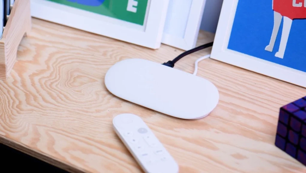 Google TV Dispositivo Google TV en color blanco sobre una mesa de madera, acompañado de un mando a distancia y un cubo tipo rompecabezas.