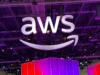 AWS invierte 50 mil millones de dólares para construir infraestructura de IA exclusiva para el gobierno de Estados Unidos Gran cartel iluminado con el logo de AWS suspendido del techo en un recinto interior, con luces moradas y estructuras metálicas visibles en el fondo.