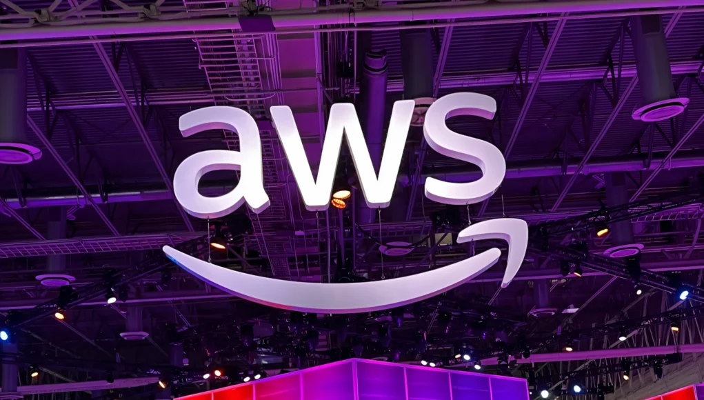 Gran cartel iluminado con el logo de AWS suspendido del techo en un recinto interior, con luces moradas y estructuras metálicas visibles en el fondo.