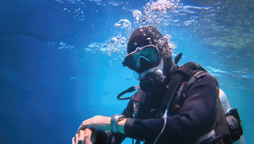 Un buzo equipado con traje de neopreno, máscara, regulador y tanque de oxígeno aparece bajo el agua, rodeado de burbujas. Está mirando o ajustando un dispositivo en su muñeca, posiblemente un ordenador de buceo, mientras nada en un entorno marino de tonos azules.