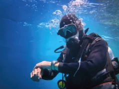 Exoesqueleto subacuático promete duplicar el tiempo de inmersión de los buzos Un buzo equipado con traje de neopreno, máscara, regulador y tanque de oxígeno aparece bajo el agua, rodeado de burbujas. Está mirando o ajustando un dispositivo en su muñeca, posiblemente un ordenador de buceo, mientras nada en un entorno marino de tonos azules.