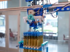 Máquina de Cornell revoluciona la fabricación 3D utilizando tejido en lugar de impresión Primer plano de un dispositivo automatizado suspendido sobre una estructura vertical llena de tubos dorados y azules. El mecanismo incluye piezas impresas en 3D, engranajes y cables electrónicos visibles, en un entorno de laboratorio luminoso con puertas de cristal y mesas al fondo.