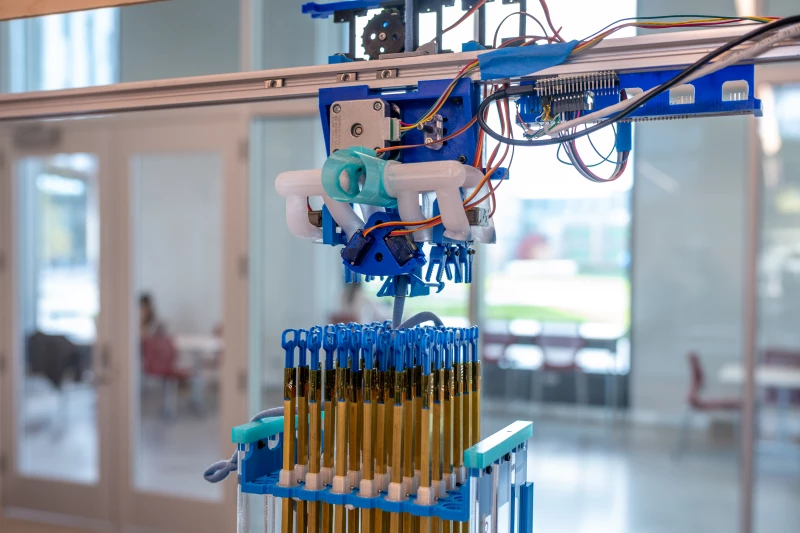 Fabricacion 3D Tejido Primer plano de un dispositivo automatizado suspendido sobre una estructura vertical llena de tubos dorados y azules. El mecanismo incluye piezas impresas en 3D, engranajes y cables electrónicos visibles, en un entorno de laboratorio luminoso con puertas de cristal y mesas al fondo.