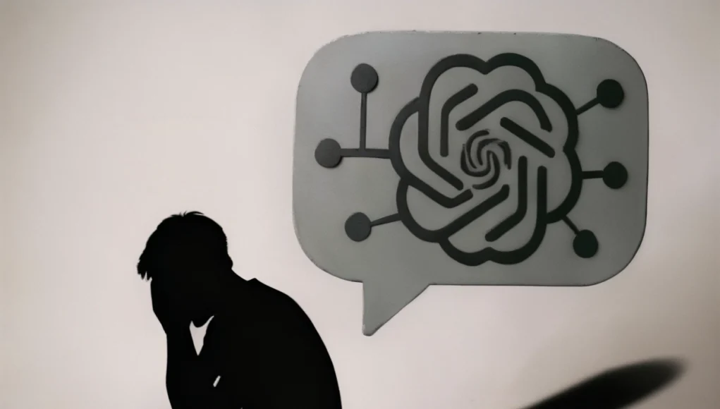IA Humanos Silueta oscura de una persona sentada en el suelo, encorvada y con la cabeza entre las manos en señal de angustia. Frente a ella aparece un gran bocadillo de conversación gris con el logotipo estilizado de una inteligencia artificial y pequeñas conexiones a su alrededor, proyectando una sombra alargada. La escena es minimalista y transmite soledad, dependencia emocional y reflexión sobre la inteligencia artificial.