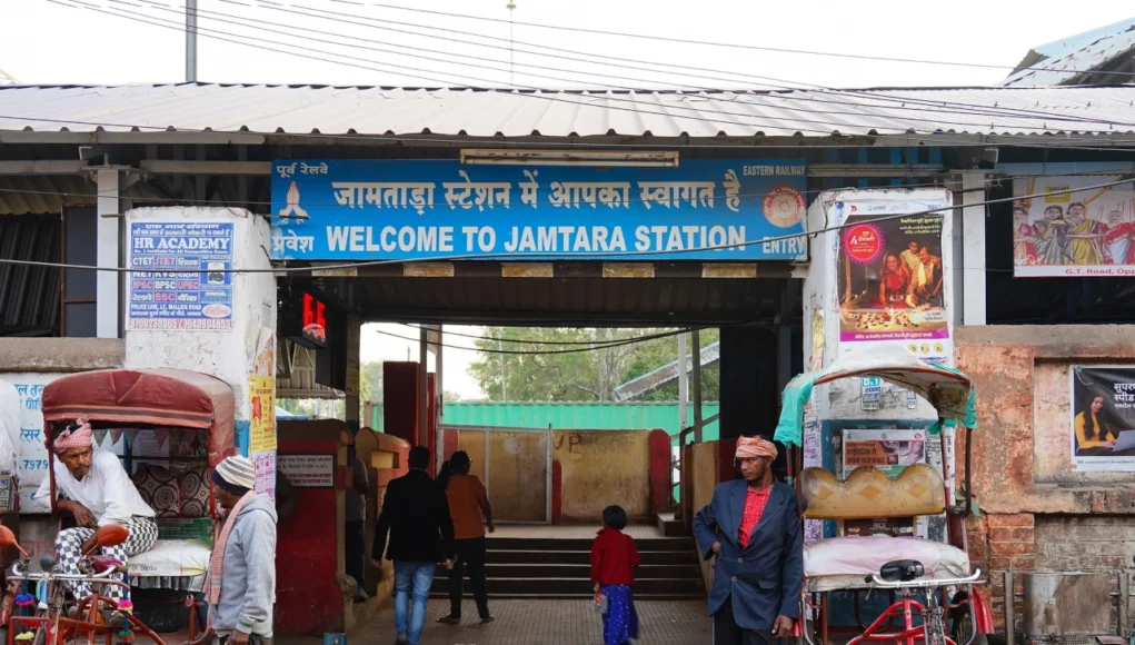 India Cibercrimen Entrada a la estación de tren de Jamtara, con un cartel que dice “Welcome to Jamtara Station” en inglés e hindi. Frente al acceso hay varias personas, rickshaws estacionados y vendedores locales.