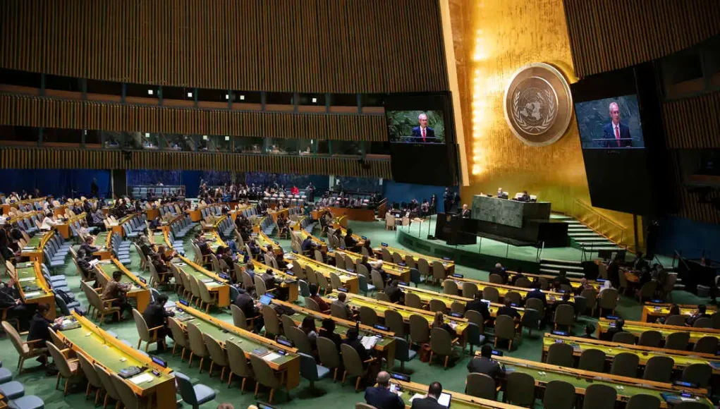Vista general del salón de la Asamblea General de las Naciones Unidas, con delegados sentados en filas curvadas mientras escuchan un discurso desde el podio central. En el fondo se observa el emblema dorado de la ONU y grandes pantallas que muestran al orador.