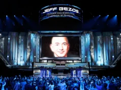 Jeff Bezos lanza una empresa de IA llamada Project Prometheus con una inversión de 6.200 millones Un gran escenario futurista iluminado en tonos azules, con un público numeroso tomando fotos. En el centro hay una enorme pantalla que muestra el rostro de un hombre calvo sonriendo ligeramente. Sobre la pantalla aparece un cartel luminoso con texto que identifica a esa persona y su cargo. En la parte inferior, varias figuras uniformadas están alineadas en formación sobre la plataforma principal.