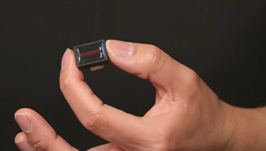 Mano sosteniendo entre los dedos un pequeño dispositivo USB negro, del tamaño de una uña. El fondo es oscuro y el enfoque está en el dispositivo y los dedos que lo sujetan.