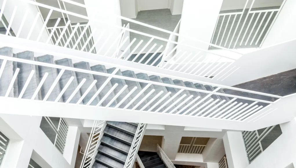 Internet Palabras Vista cenital de un interior arquitectónico moderno con múltiples tramos de escaleras blancas y barandillas geométricas que se cruzan en distintos niveles, creando un patrón abstracto y simétrico de líneas y formas.