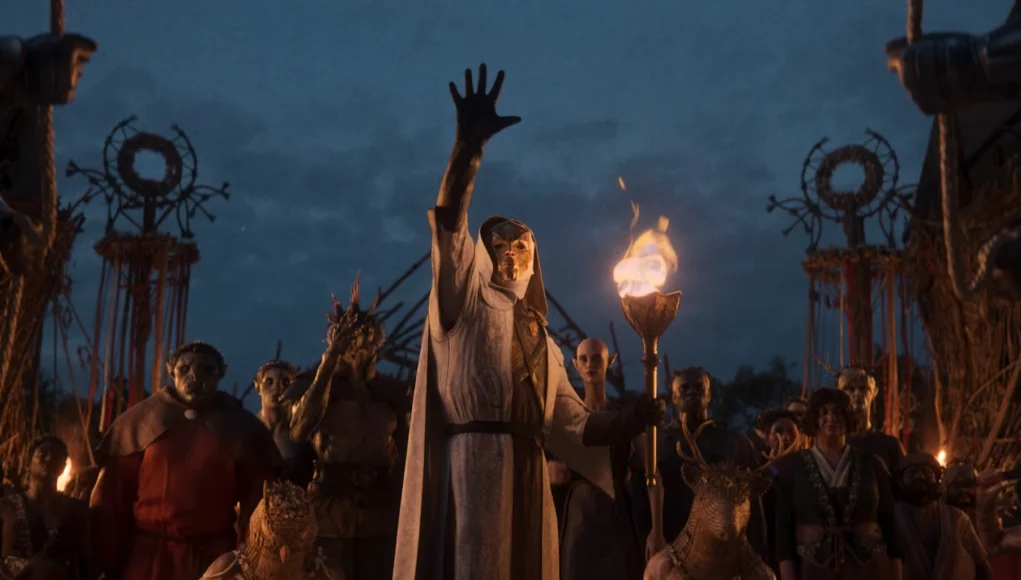 Figura central con túnica clara y máscara ritual alzando una mano mientras sostiene una antorcha encendida, rodeada por un grupo de personajes de aspecto fantástico y criaturas humanoides en un ritual nocturno, bajo un cielo oscuro con estandartes y símbolos tribales al fondo.