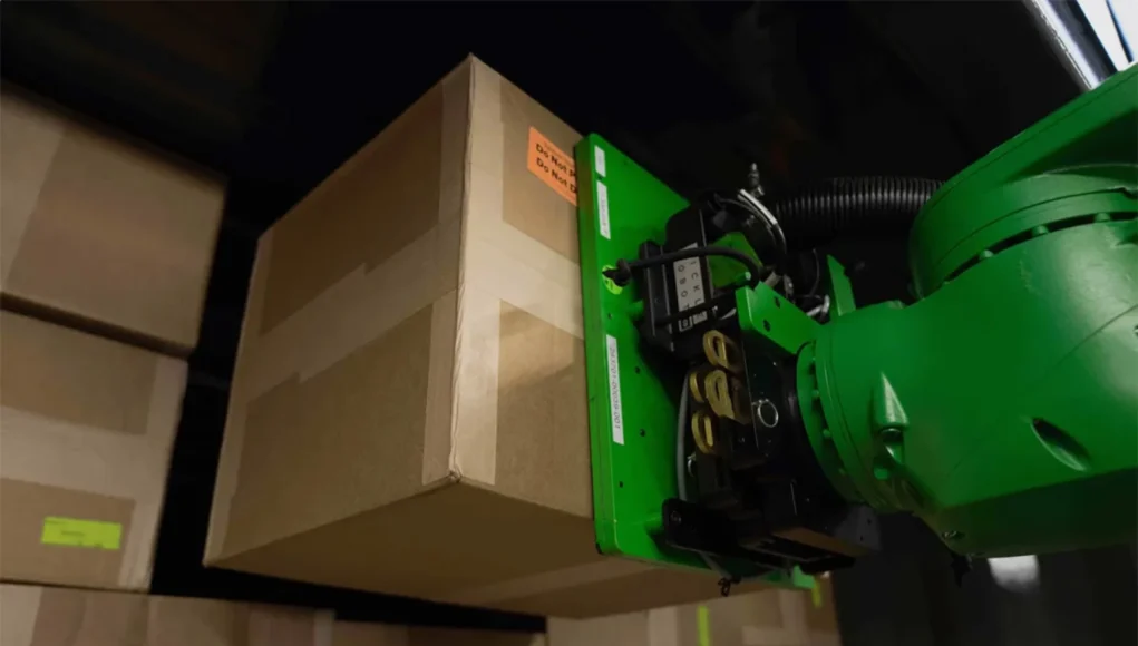MIT Robot Un brazo robótico de color verde levanta una caja de cartón en un almacén lleno de paquetes apilados. El robot sostiene la caja mediante un mecanismo de agarre en su extremo.