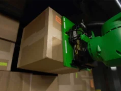 MIT desarrolla un robot industrial que descarga 34 toneladas de carga por hora con succión neumática Un brazo robótico de color verde levanta una caja de cartón en un almacén lleno de paquetes apilados. El robot sostiene la caja mediante un mecanismo de agarre en su extremo.