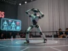 Tesla Optimus cae en demostración de Miami, movimientos de manos generan debate sobre operación remota Un robot humanoide metálico realiza una pose con las manos sobre la cabeza en un escenario circular durante una demostración tecnológica. Detrás se observa una pantalla digital con diagramas del robot y varias personas sentadas como público. Botellas de agua están distribuidas en el borde del escenario, y cortinas grises forman el fondo.