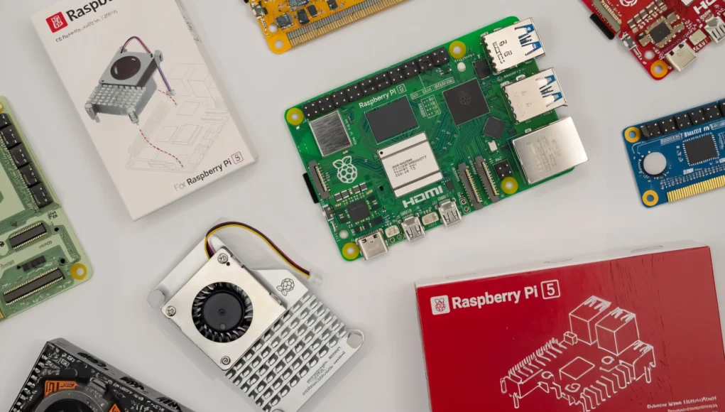 Raspberry IA Varios componentes electrónicos dispuestos sobre una superficie clara, incluyendo una placa Raspberry Pi 5, cajas de embalaje, una unidad de refrigeración con ventilador y otras placas de expansión y accesorios.