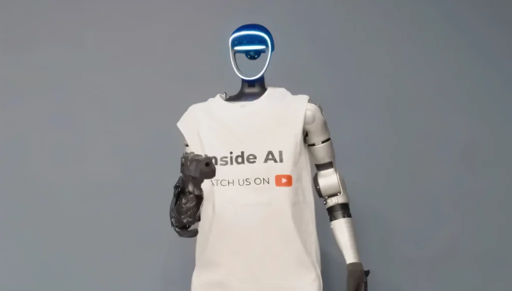 Robot Seguridad Robot humanoide de aspecto futurista con rostro iluminado en azul, vestido con una camiseta blanca con el texto “Inside AI” y un icono de YouTube, de pie frente a un fondo neutro gris, representando la divulgación y presencia de la inteligencia artificial en medios digitales.
