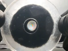 Los robots blandos ya pueden ver con un ojo artificial autoenfocable que imita al humano Primer plano de un disco transparente y flexible sostenido con guantes negros; en el centro se observa una pequeña estructura multicolor brillante, similar a un objeto holográfico o un marcador óptico.