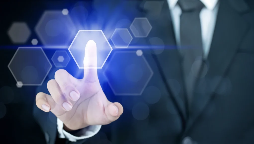 Pantalla Sentidos Mano de una persona con traje tocando una interfaz digital futurista, con un dedo presionando un icono hexagonal luminoso en tonos azules que representa autenticación o acceso tecnológico.
