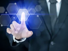 Investigadores de UCSB desarrollan pantalla táctil que permite sentir las imágenes Mano de una persona con traje tocando una interfaz digital futurista, con un dedo presionando un icono hexagonal luminoso en tonos azules que representa autenticación o acceso tecnológico.