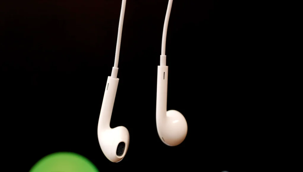Auriculares blancos con cable suspendidos frente al logotipo de Spotify desenfocado sobre fondo oscuro, evocando el consumo de música y audio en plataformas digitales.