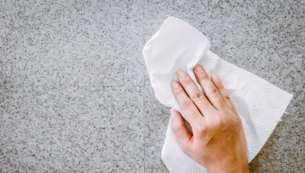 Escudo Auditivo TDA Mano limpiando una superficie de piedra o encimera gris con una servilleta de papel blanca, representando una acción de limpieza doméstica.